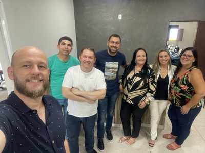 na foto temos sete pessoas em uma sala com parede de fundo cinza. A primeira pessoa, no canto esquerdo, é um homem branco de cerca de quarenta anos de idade. Ele segura o celular para tirar a foto. Ao seu lado, um homem de certa de quarenta anos vestindo blusa verde. No centro um home de cerca de cinquenta anos, vestindo branco e de braços cruzados, ao seu lado outro homem com blusa preta, aparentando ter quarenta anos. Ao lado deste, uma senhora de cerca de cinquenta anos, com blusa estampada em preto e calça preta. No canto direito, duas mulheres, uma com roupas brancas, aparentando ter cinquenta anos e uma com roupa estampada colorida e calça preta, com aparência de trinta e cinco anos. todas as pessoas estão de pé e em fila
