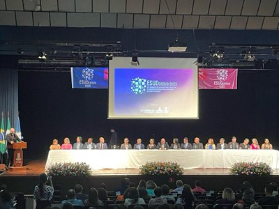 Imagem inicial do evento ESUD com o banner do evento ao fundo, pessoas sentadas na plateia e uma mesa grande em cima do palco com vários representantes do evento
